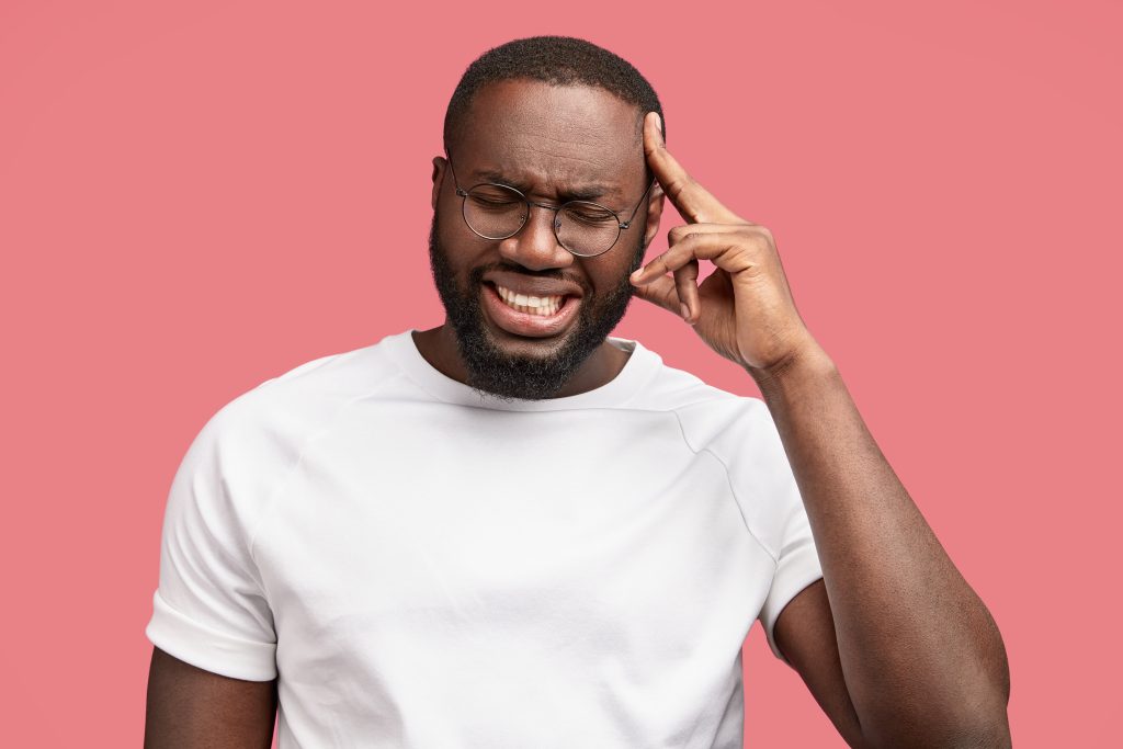 stressed dark skinned male entrepreneur faces financial crisis, has unhappy look, keeps fore finger on head, clenches teeth, closes eyes, finds solution in mind, being in panic needs help from someone