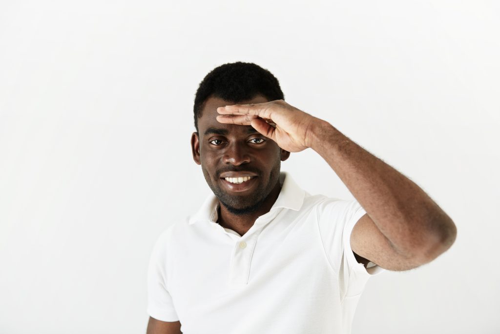 close up portrait of young curious african american man peering ahead into the distance, with hand on his forehead, searching for something or looking to the future at the camera with dreamy expression
