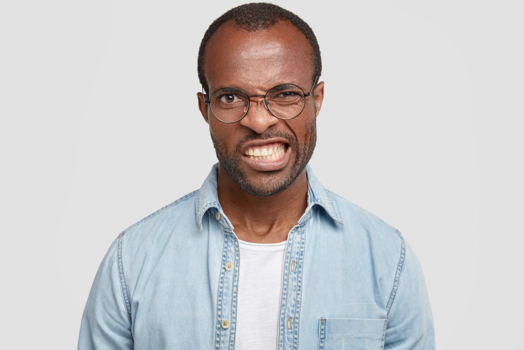 irritated african american guy frowns face in displeasure, being annoyed with bad news, clenches teeth, wears fashionable denim shirt and glasses, isolated over white background. negative emotions